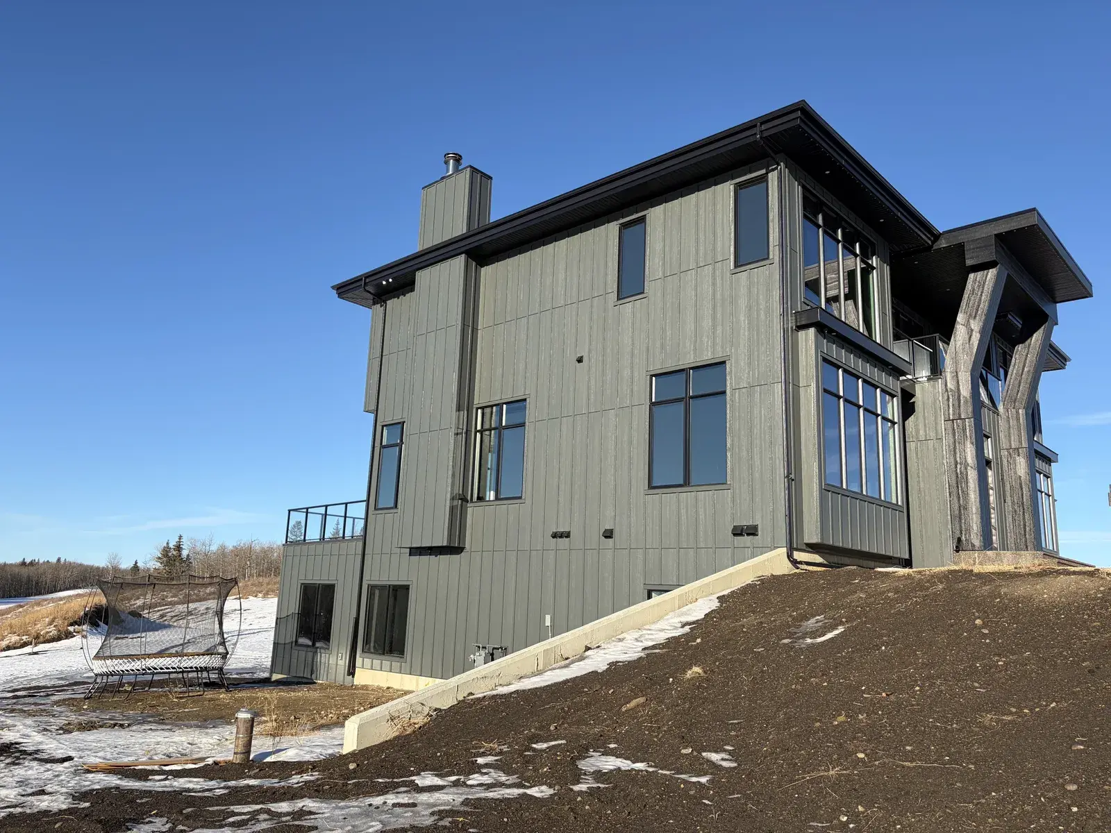 Side elevation of a Lacombe-area custom home in grey-green vertical board-and-batten siding with black trim and large windows, finished by PJM