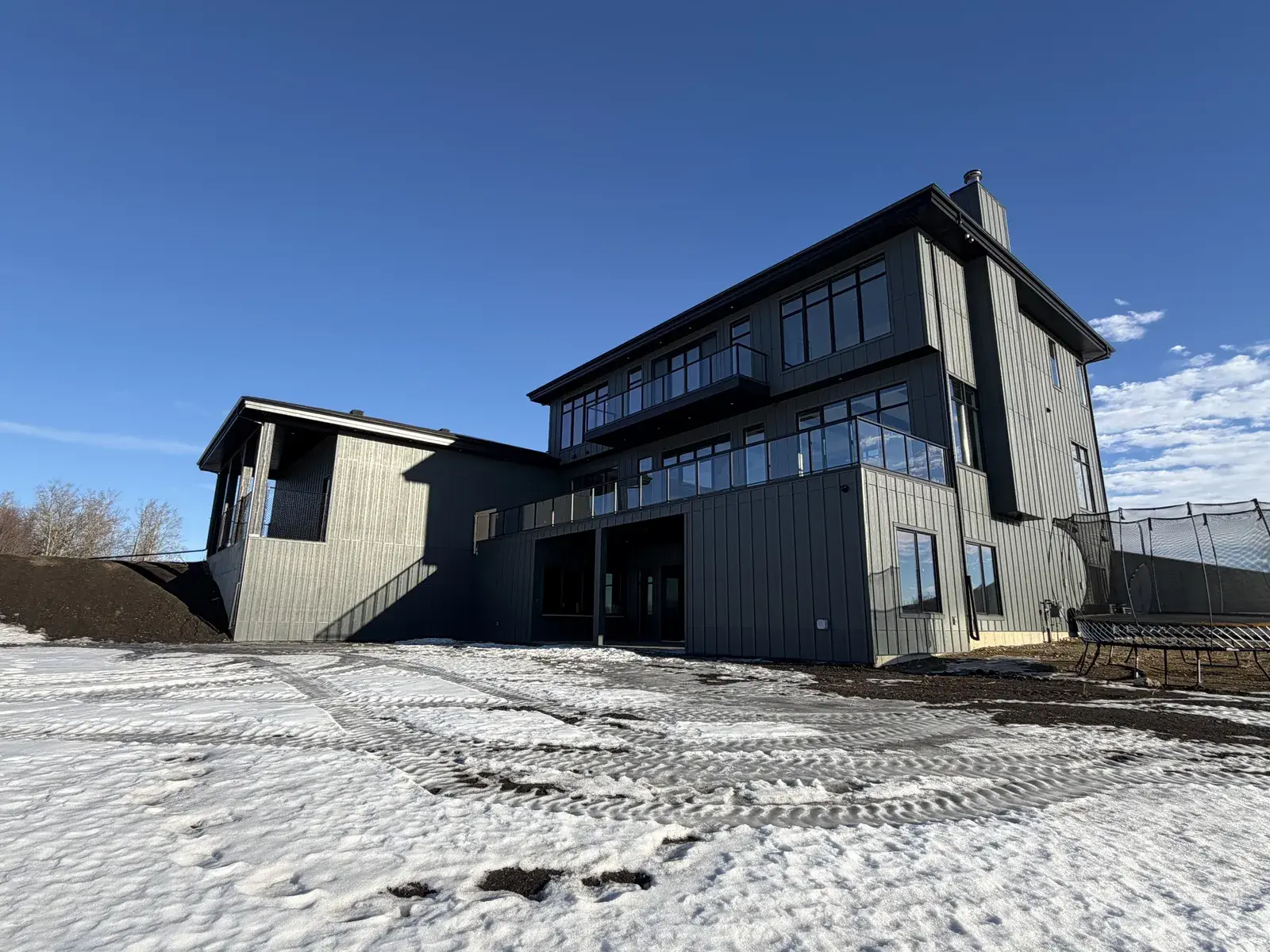 Rear of a tall walk-out custom home near Lacombe in dark board-and-batten siding with a covered upper deck and full-height black-framed glazing by PJM