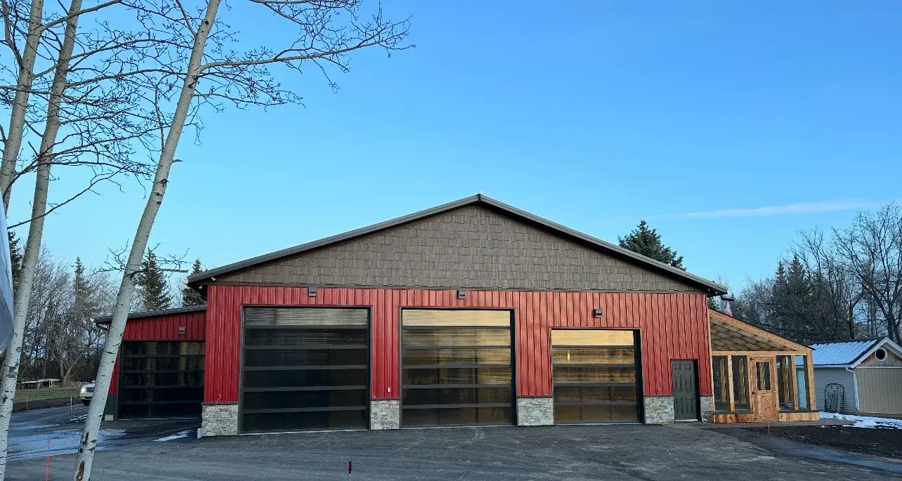 Red board-and-batten shop with a shake gable, glass overhead doors and a stone base in Red Deer County, finished by PJM Home Advantage