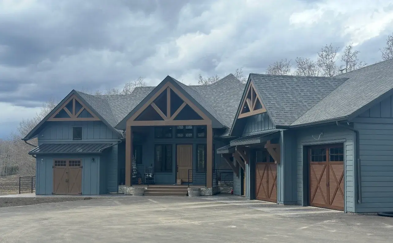 Blue-grey timber-frame acreage home in Red Deer County with board-and-batten and lap siding, wood carriage garage doors and stone accents, finished by PJM Home Advantage