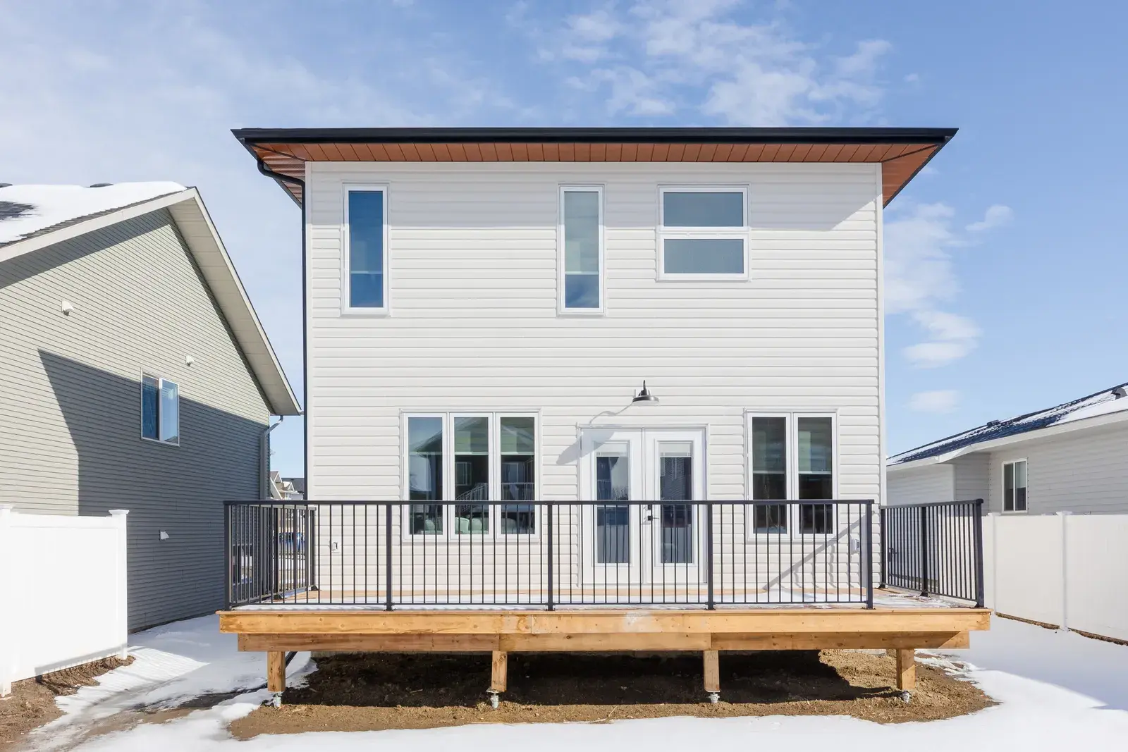 Rear elevation of a Blackfalds two-storey in white lap siding with a wood deck, French doors and dark soffit and fascia by PJM